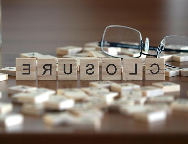 Scrabble tiles spelling CLOSURE on the tile rack with other tiles scattered on a table with eyeglasses.